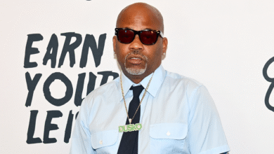 Damon Dash wearing sunglasses, a light blue shirt, and a necklace with "Dusko" pendant, stands in front of a white background.