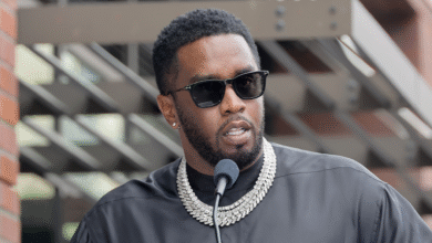 Sean "Diddy" Combs speaking at a podium, wearing sunglasses and a black outfit with a diamond necklace.