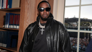 Diddy in a black leather jacket and sunglasses stands indoors, with a crowd visible through the window behind him.