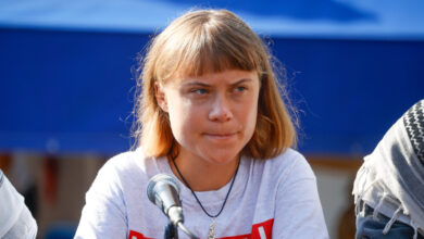 Greta Thunberg, wearing a white shirt, sits at a table with a microphone, looking thoughtful against a blue background.