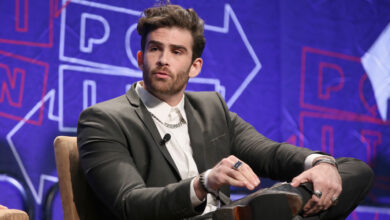 Hasan in a suit sits on stage, speaking into a microphone, with a backdrop featuring arrows and text.