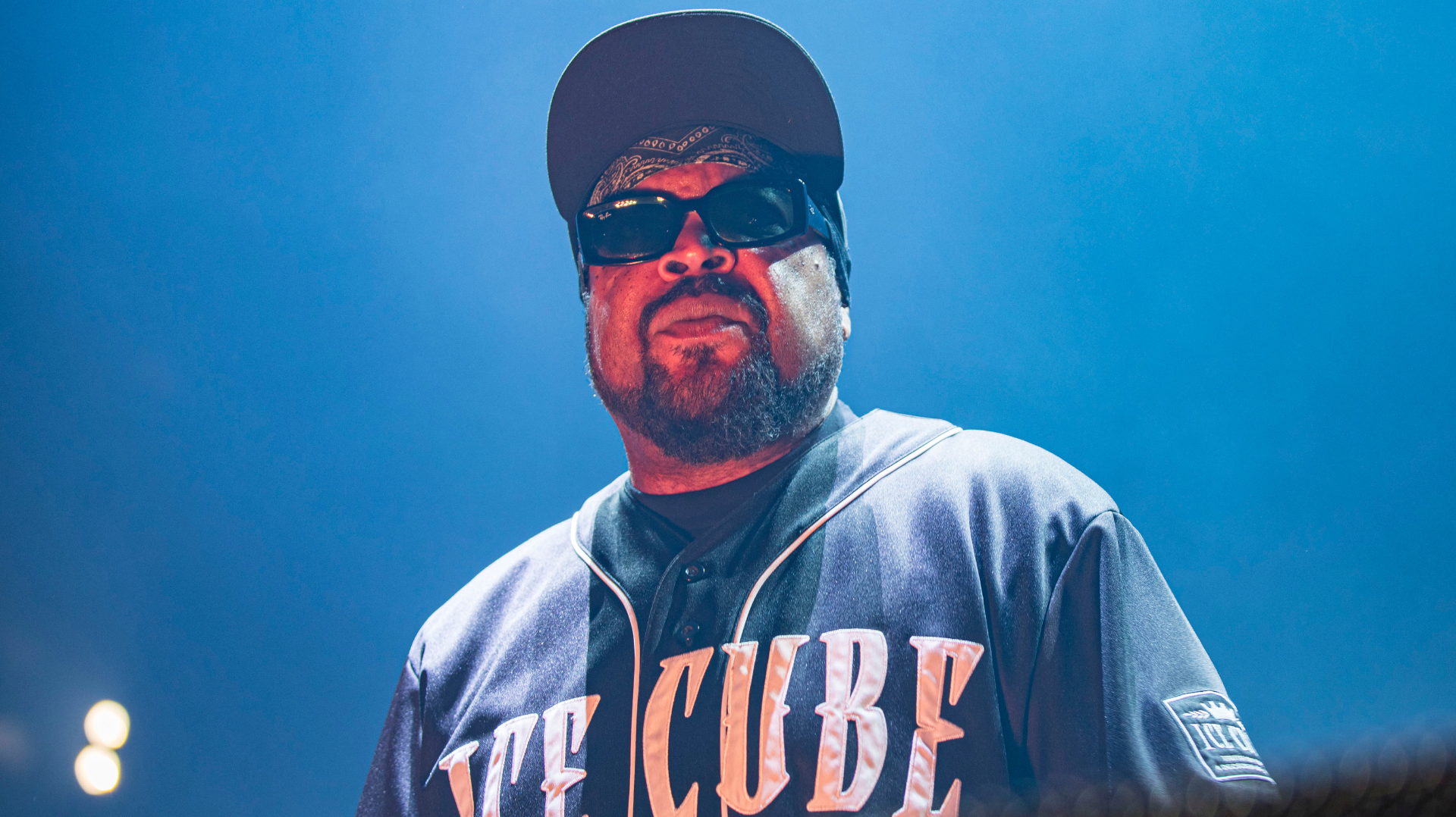 Ice Cube’s Tour Bus Dammaged in Fire Was ‘Random Act of V... 1 Ice Cube on stage wearing sunglasses and a cap, with a serious expression, under blue lighting.
