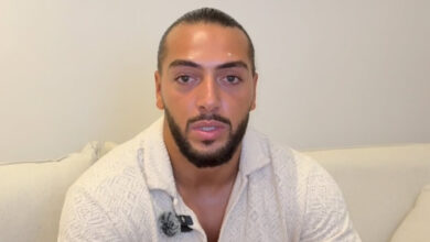 French Influencer Amine Mojito with a beard and slicked-back hair sits on a couch, wearing a textured white shirt with a microphone attached.