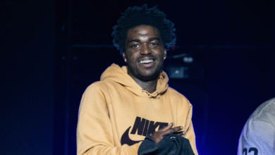 A man with short curly hair wearing a yellow Nike hoodie smiles on stage.
