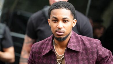 DDG wears a burgundy jacket featuring a subtle, intricate pattern. The jacket has a pointed collar and a zip fastening. Underneath, a brown ribbed shirt is visible. Jewelry includes multiple layered necklaces with a prominent pendant and small stud earrings. Hair is styled in a short, textured cut, outside Amiri, during Paris Fashion Week - Menswear Spring/Summer 2026, on June 26, 2025 in Paris, France.